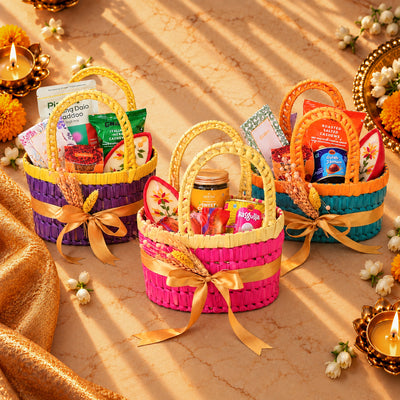 Three colorful woven gift baskets filled with assorted packaged snacks, tea, and sweets, arranged on a warm beige surface with decorative candles, flowers, and draped fabric.
